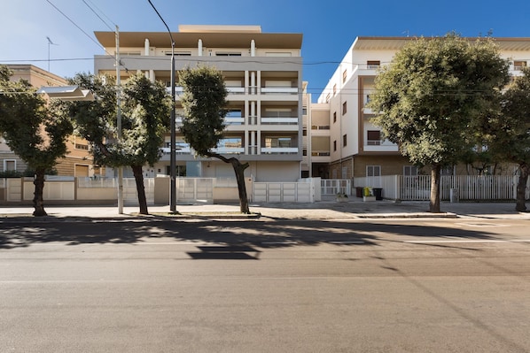 A multi-story residential building with balconies, a gated entrance, and trees lining the street.