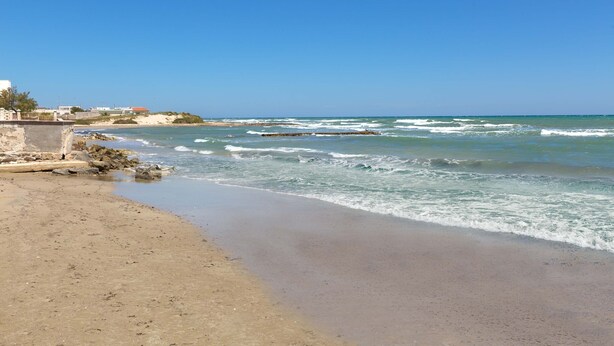 A beach with a rocky shoreline, waves, and buildings in the background. - 1939 Villetta Camilla (Lecce)
