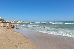 A beach with a rocky shoreline, waves, and buildings in the background. - 1939 Villetta Camilla (Lecce)
