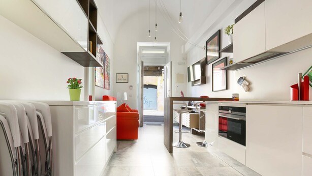 A modern kitchen with white cabinets, a red chair, and a dining area with a table and chairs. - 1991 Suite Verdi (Lecce)