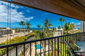 A balcony with a view of a swimming pool, palm trees, and a distant ocean. - Kihei Akahi #D-513 Oceanview, Across from Kamaole Beach ll, Great Rates! (Kihei)