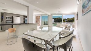 Una cocina moderna con una isla central, un comedor con mesa de vidrio y vistas al exterior.