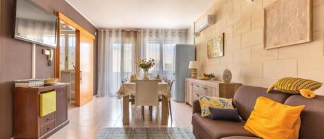 Una sala de estar moderna con un sofá, una mesa de comedor preparada para cuatro personas, un televisor montado en la pared y una cocina visible al fondo.