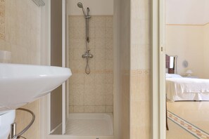 A bathroom with a shower, a sink, and a bed in the adjacent room.