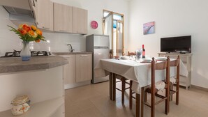 A compact kitchen with a gas stove, a refrigerator, and a dining area with a table set for two. - 1826 Villetta Solaire (Porto Cesareo)