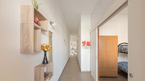 A narrow hallway with a wooden wardrobe, a small shelf with a vase of flowers, and a bed with a floral bedspread. - 1826 Villetta Solaire (Porto Cesareo)