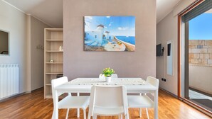 Uma área de refeições com mesa e cadeiras brancas, um quadro com uma paisagem costeira e vista através de uma porta de vidro.