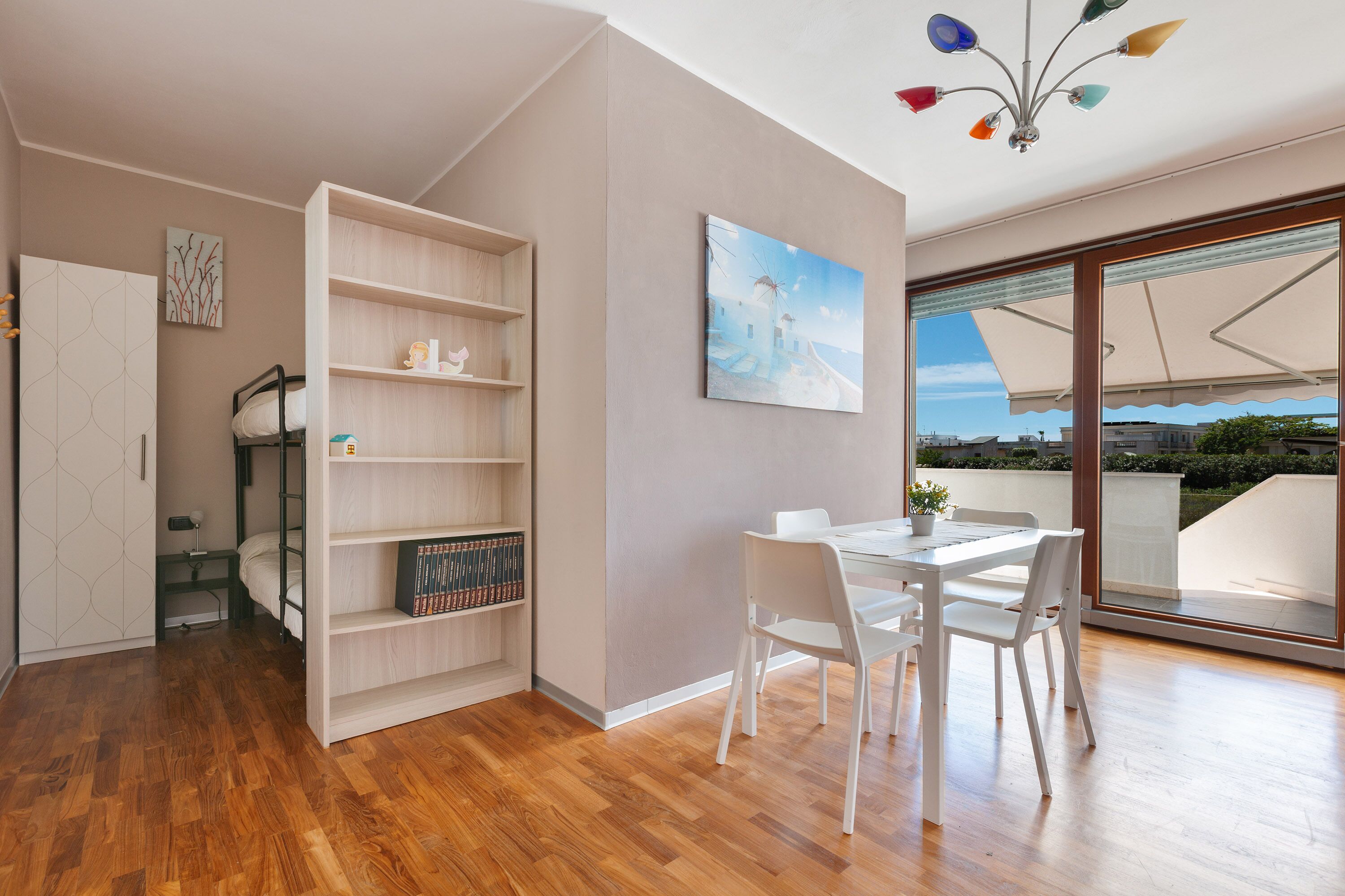 A modern room with a wooden floor, a white dining table with chairs, a bookshelf, a bed with a ladder, and a large window with a view of buildings and greenery.