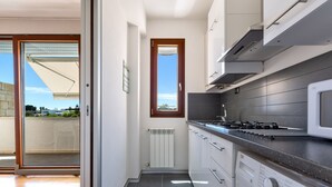 Cozinha moderna com fogão embutido, micro-ondas e máquina de lavar roupa, porta de vidro deslizante para uma varanda e vista para a paisagem urbana.