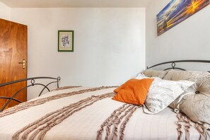 A neatly made bed with a ruffled bedspread and a single orange pillow, a framed botanical print on the wall, and a door to the left.