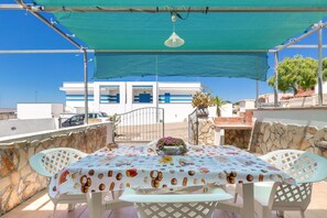 An outdoor patio with a table set for two, covered by a blue shade, and a view of a white building with blue accents in the background.
