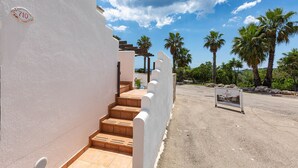 Une entrée d’immeuble avec un panneau indiquant le numéro, un escalier et une enseigne, le tout dans un environnement extérieur ensoleillé.