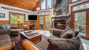 A cozy living room with a stone fireplace, wooden ceiling, and large windows.
