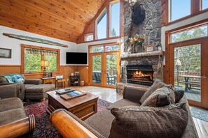A cozy living room with a stone fireplace, wooden ceiling, and large windows.