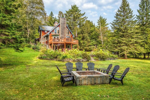 A log cabin with a deck and a fire pit surrounded by Adirondack chairs. - Brooklake - Custom built home on Dodge Pond, with 250' on Dodge Pond Stream too (Rangeley)