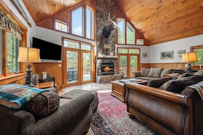 A spacious living room with a stone fireplace, wooden furniture, and large windows. - Brooklake - Custom built home on Dodge Pond, with 250' on Dodge Pond Stream too (Rangeley)