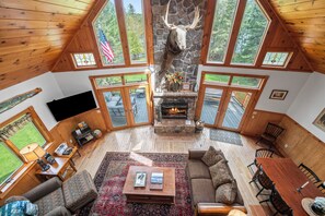 A spacious living area with a stone fireplace, wooden ceiling, and large windows. - Brooklake - Custom built home on Dodge Pond, with 250' on Dodge Pond Stream too (Rangeley)