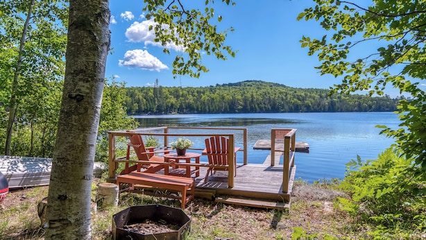 A lakeside deck with wooden furniture and a fire pit, overlooking a calm lake and forested hills.