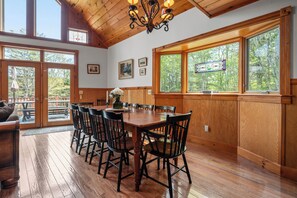 A dining area with a wooden table and chairs, large windows with a view of trees, and a chandelier.