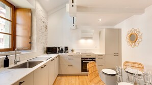 Una cocina moderna con suelo de madera, armarios blancos, microondas integrado y comedor con sillas de mimbre y una mesa de vidrio.