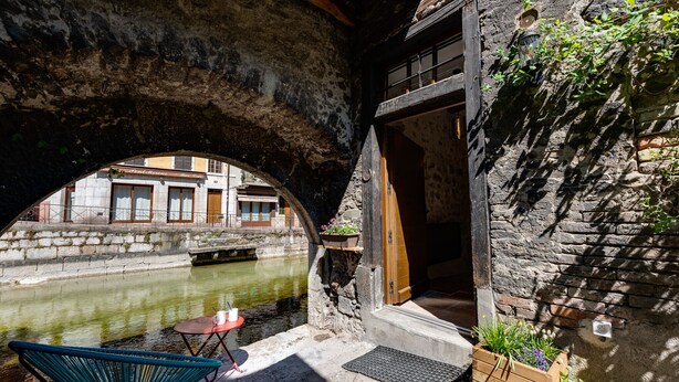 Un área de estar al aire libre con una silla y una mesa, un muro de piedra con una puerta de madera, y un canal con edificios al fondo.