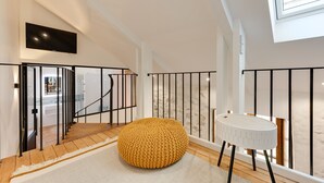Un interior moderno con una escalera, un reposapiés amarillo redondo, una mesita blanca y un televisor montado en la pared.
