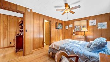 A bedroom with wooden paneling, a bed with a blue cover, a wooden chair, a bedside table with a lamp, and a ceiling fan.