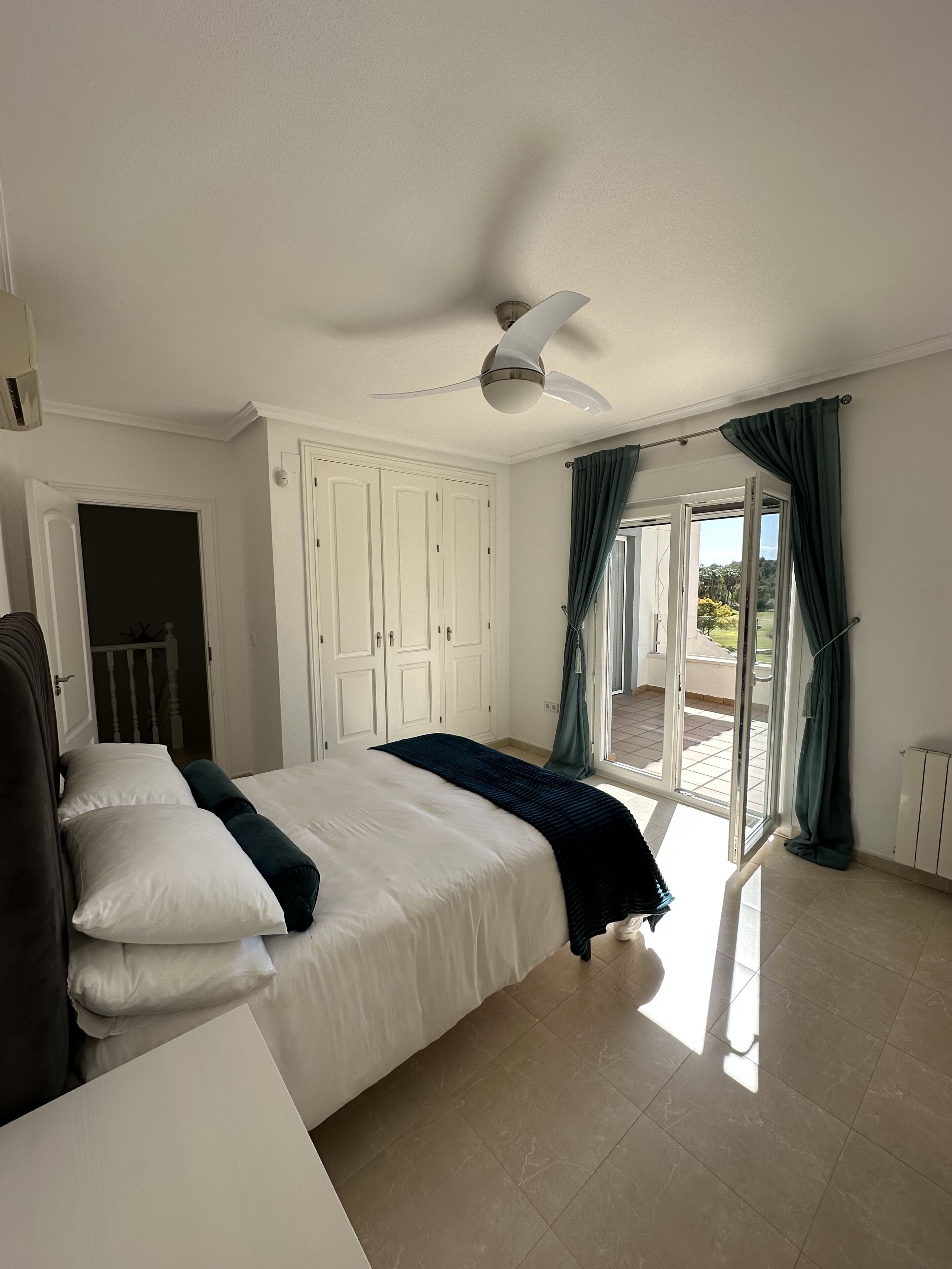 A bedroom with a bed, a ceiling fan, a sliding glass door to a balcony, and a white wardrobe.