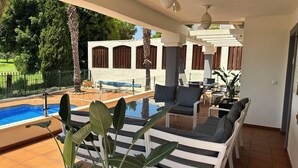 A covered outdoor area with a pool, lounge chairs, and a view of a white building with wooden shutters. -  Luxury Front-Line Golf Villa with Private Pool & Panoramic Views (Orihuela)