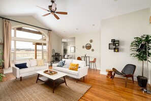 A spacious living room with a white sofa, a coffee table, and a ceiling fan. There is a dining area with a table and chairs, and a kitchen in the background.