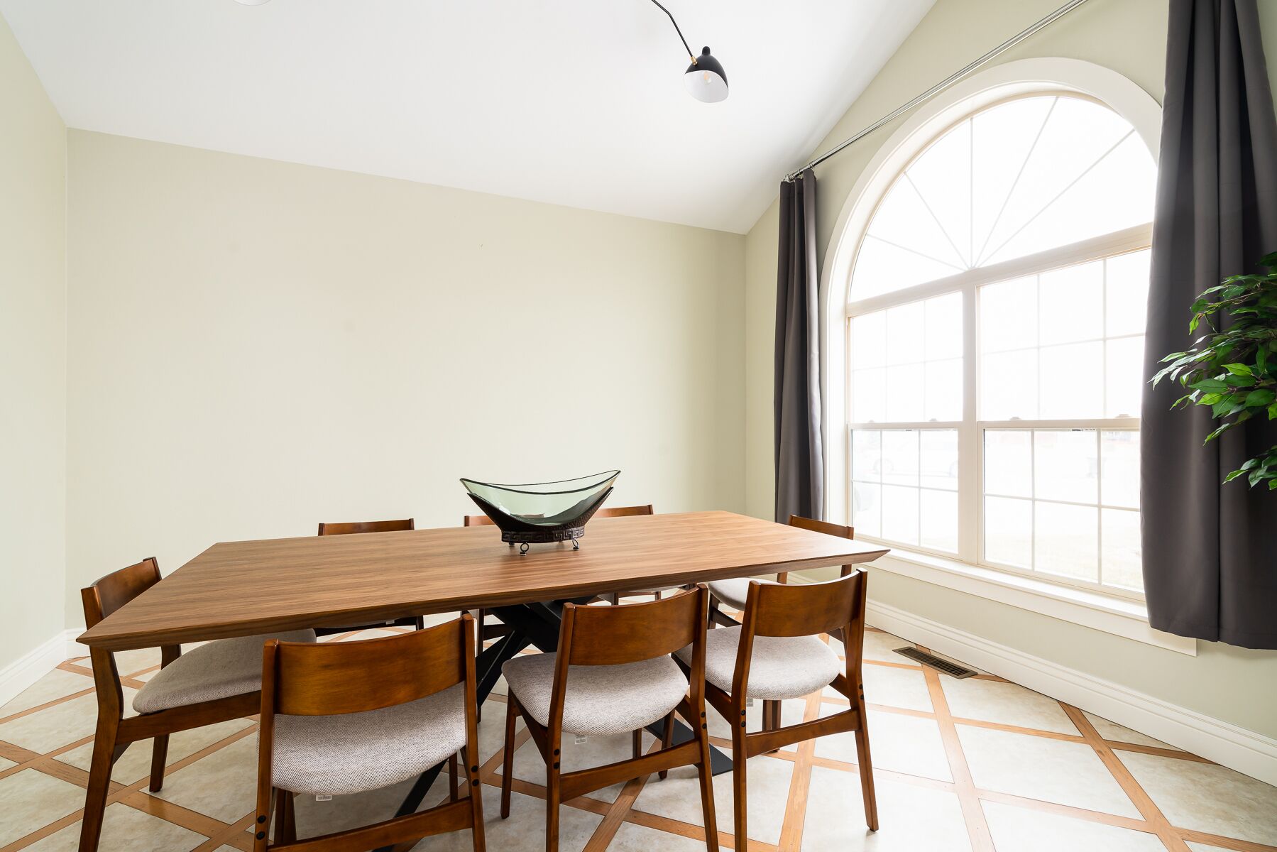 Un coin repas avec une table en bois, six chaises, un centre de table composé d’un vase en verre et une grande fenêtre agrémentée de rideaux.