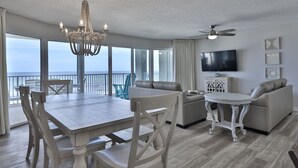 A spacious living area with a dining table, sofas, and a chandelier. A television is mounted on the wall, and there is a balcony with a view of the ocean.