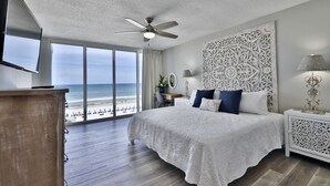 A bedroom with a large bed, a ceiling fan, a TV, and a view of the beach.