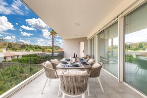 Un comedor con una mesa lista para cuatro personas, rodeado de sillas de mimbre, con vistas a un paisaje urbano y vegetación.
