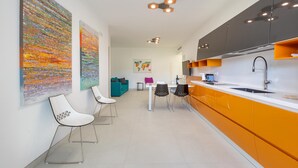 Une cuisine moderne avec des meubles de couleur orange, un plan de travail blanc et un évier noir. On y trouve deux chaises blanches avec des assises noires, ainsi qu’une petite table sur laquelle repose un coussin rose, le tout sur un fond neutre.