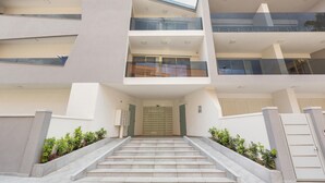 Un immeuble moderne à plusieurs étages, doté d’une entrée centrale, d’un escalier menant à l’entrée et d’un balcon en verre situé à l’étage supérieur.