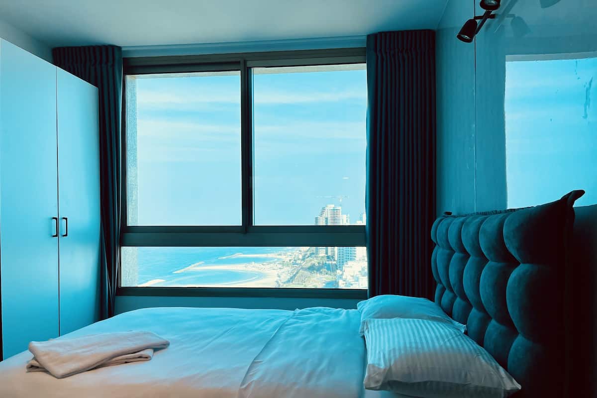 A bedroom with a large window, a bed with white bedding, and a view of a beach and buildings.
