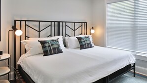 Una cama bien tendida con ropa blanca y dos almohadones decorativos, un cabecero de metal negro y dos lámparas de lectura fijadas a la pared.