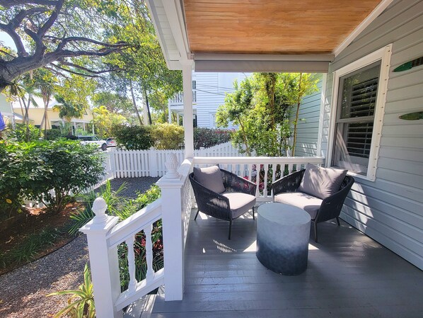 A covered porch with two chairs and a round table, surrounded by a white picket fence and greenery.