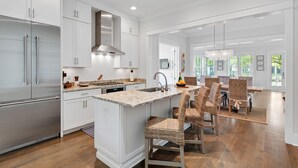 A modern kitchen with white cabinetry, a central island, and a dining area with wicker chairs. - WaterColor on Dragonfly Park | Porches & Park Views (Santa Rosa Beach)