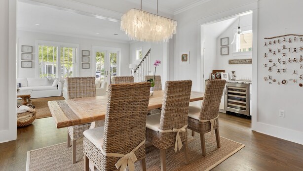 A dining area with a wooden table and wicker chairs, a chandelier, and a kitchen in the background. - WaterColor on Dragonfly Park | Porches & Park Views (Santa Rosa Beach)