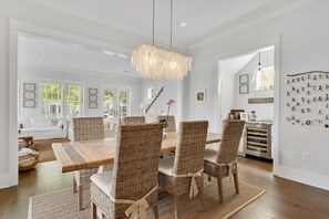 A dining area with a wooden table and wicker chairs, a chandelier, and a kitchen in the background. - WaterColor on Dragonfly Park | Porches & Park Views (Santa Rosa Beach)