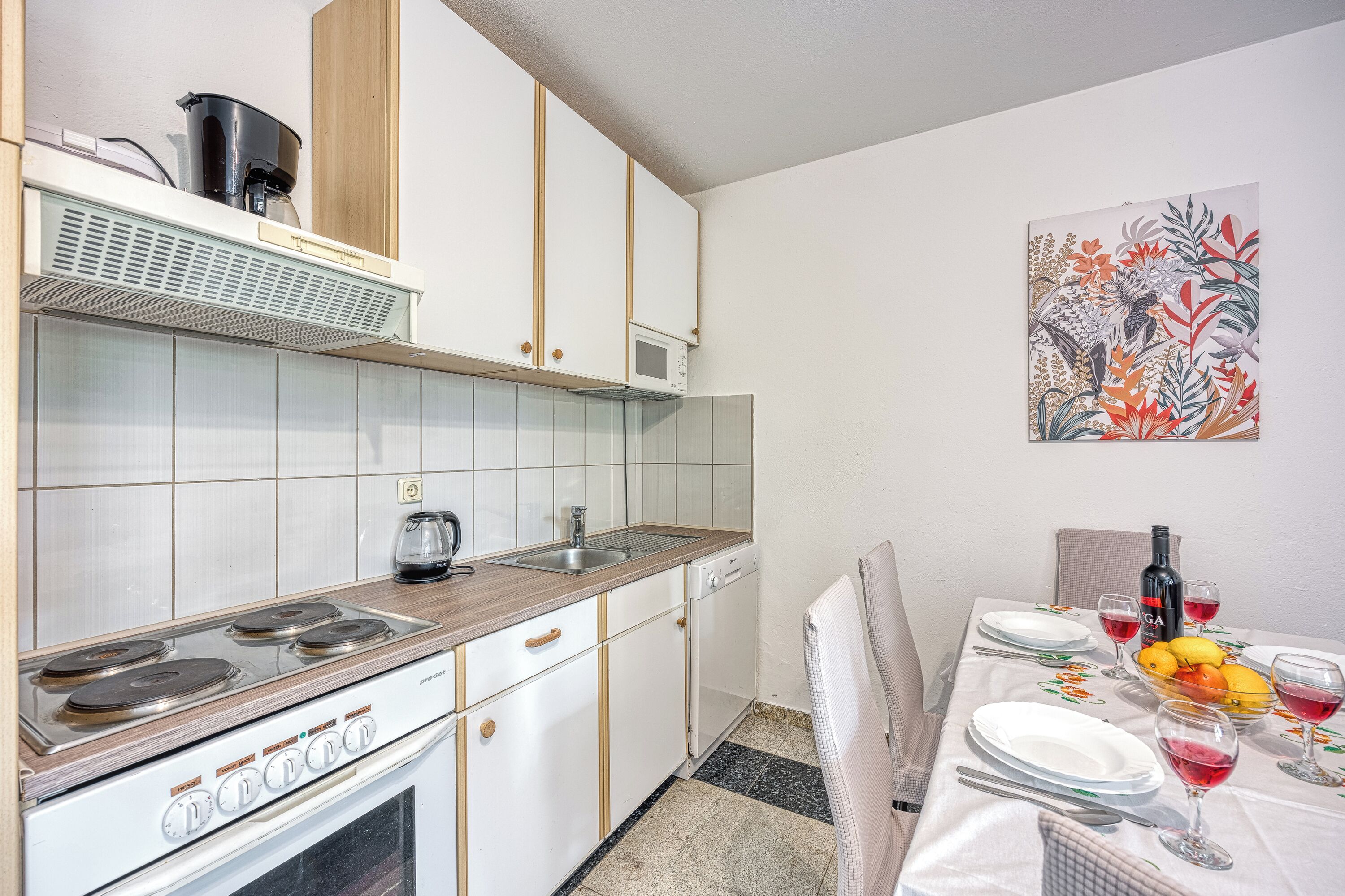 Una cocina con estufa, lavaplatos y armarios, una mesa de comedor con platos y vasos, y un cuadro en la pared.