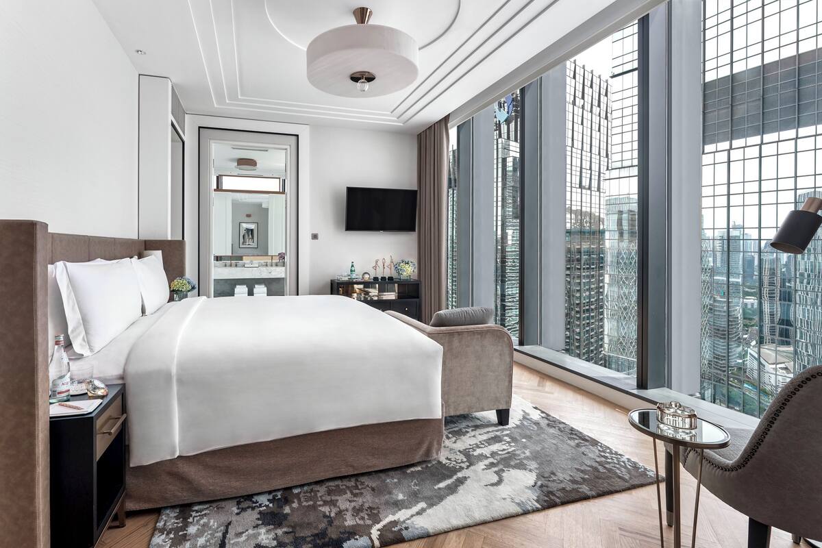 Quarto de hotel moderno com uma cama grande, uma escrivaninha pequena, uma cadeira e uma janela ampla com vista para a cidade.