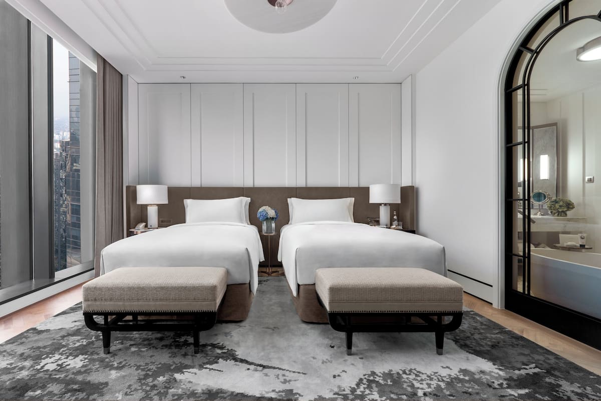 Quarto de hotel moderno com duas camas, uma janela grande com vista para a cidade, um banheiro com espelho e uma pequena mesa com luminária.