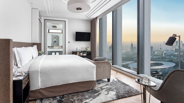 A modern hotel room with a large bed, a desk, a chair, a small table, and a view of a cityscape through large windows. - The Langham, Jakarta (Jakarta)