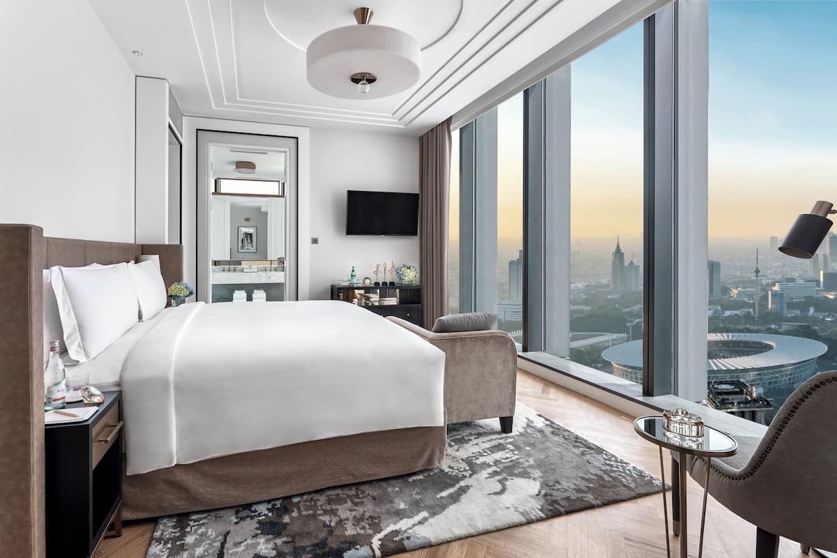 Quarto de hotel moderno com uma cama grande, uma escrivaninha, uma cadeira, uma mesinha de centro e vista para a paisagem urbana pelas amplas janelas.