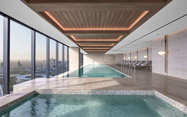 A modern indoor pool with a city view, wooden ceiling, and lounge chairs along the side. - The Langham, Jakarta (Jakarta)