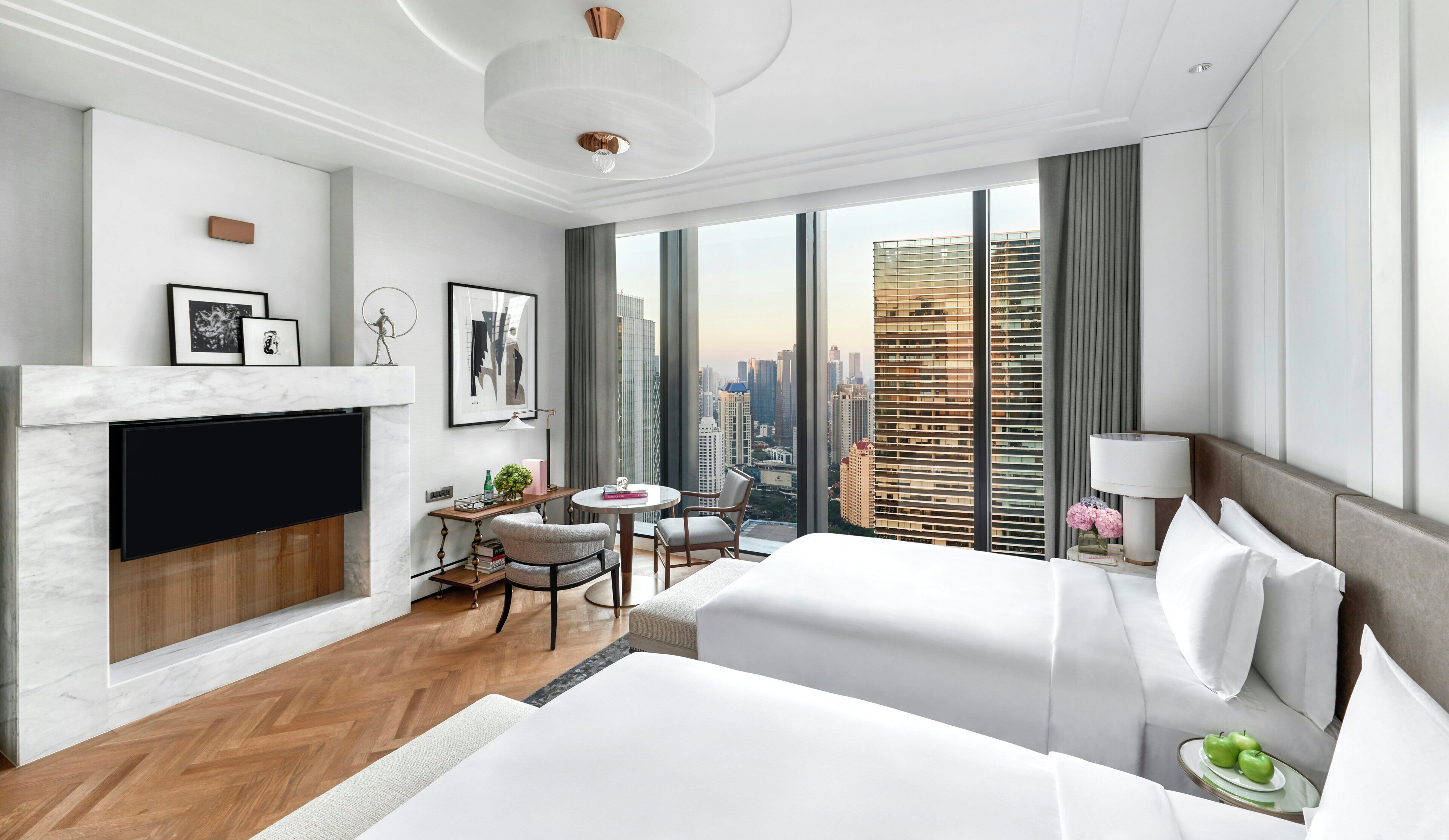 A modern hotel room with a large bed, a desk with a chair, a small table, a TV mounted on the wall, and a view of the cityscape through large windows.