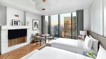 A modern hotel room with a large bed, a desk with a chair, a small table, a TV mounted on the wall, and a view of the cityscape through large windows.